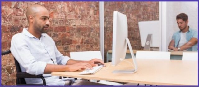 A man in a wheelchair sits at a desk in an office environment and uses a computer. 