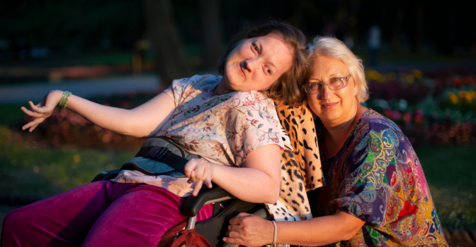 Young woman in wheelchair with her older carer by her side, outside in the sun.