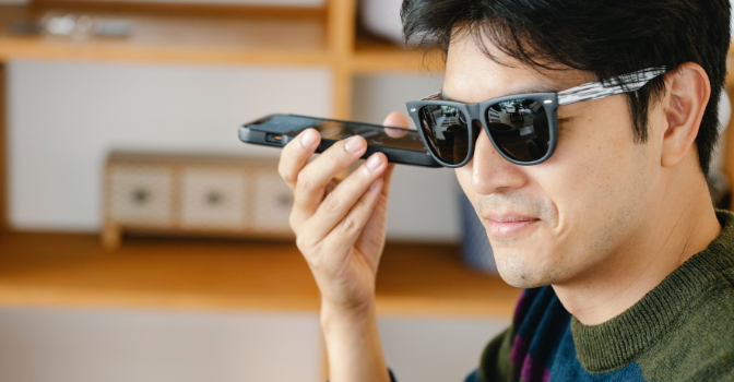 A vision impaired man is sittin wearing sunglasses and holding his phone to his ear.